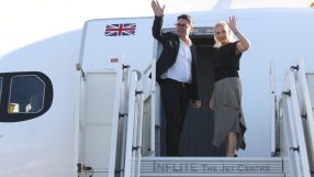 Andy Burnham and a woman waving from the steps of an airplane with a Union Jack flag