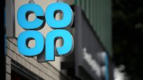 Close-up of a blue illuminated Co-op sign hanging outside a building, with a dark background.