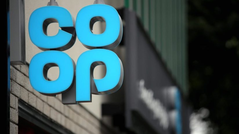 Close-up of a blue illuminated Co-op sign hanging outside a building, with a dark background.
