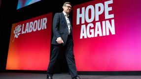 A man in a suit walking across a stage with LABOUR and HOPE AGAIN displayed on a red and pink screen behind him.