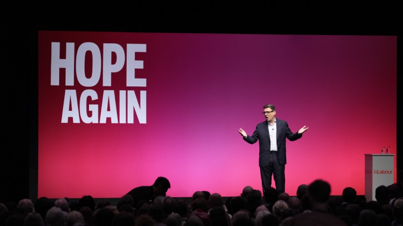 Man in suit speaking on stage in front of a large screen displaying HOPE AGAIN to an audience, likely at a political or bu...