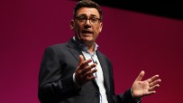 Andy Burnham, Mayor of Greater Manchester, speaking at a podium with hands gesturing, against a red background.