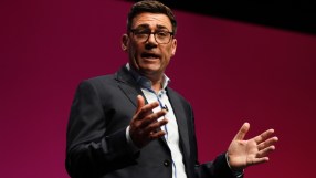 Andy Burnham, Mayor of Greater Manchester, speaking at a podium with hands gesturing, against a red background.