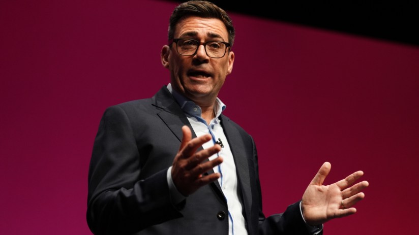 Andy Burnham, Mayor of Greater Manchester, speaking at a podium with hands gesturing, against a red background.