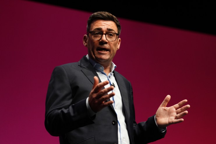 Andy Burnham, Mayor of Greater Manchester, speaking at a podium with hands gesturing, against a red background.