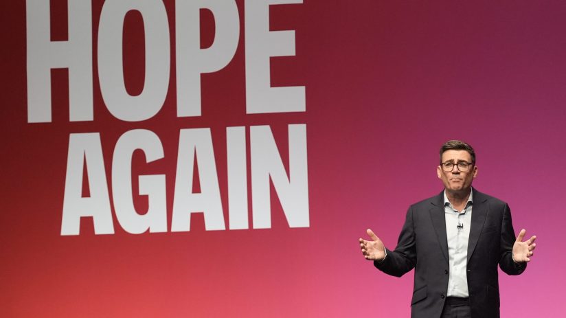 Andy Burnham speaking at a podium with HOPE AGAIN displayed prominently behind him, gesturing with hands.