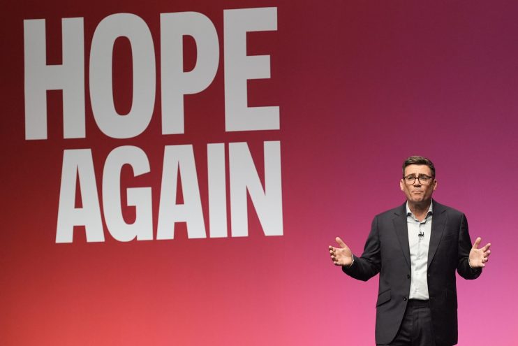 Andy Burnham speaking at a podium with HOPE AGAIN displayed prominently behind him, gesturing with hands.