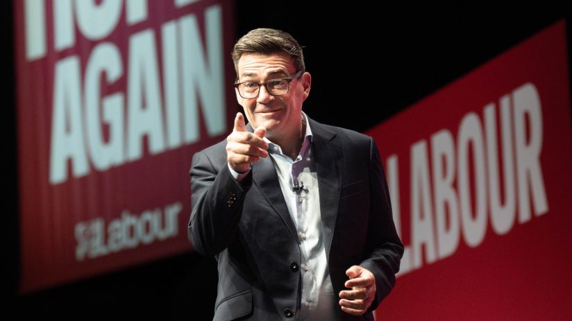 Andy Burnham, wearing glasses and a suit, points forward with a Labour banner in the background.
