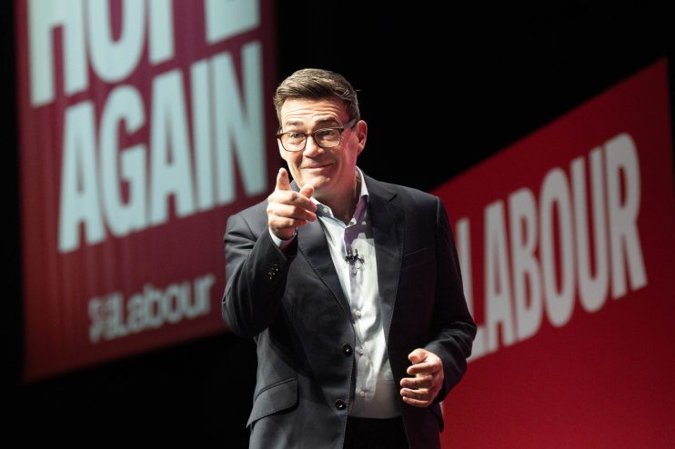 Andy Burnham, wearing glasses and a suit, points forward with a Labour banner in the background.