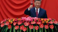 Xi Jinping in a dark suit and red tie, raising a glass of red wine, with a floral arrangement below him.