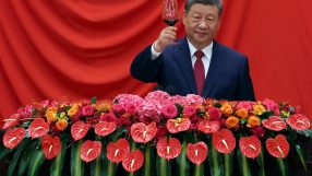 Xi Jinping in a dark suit and red tie, raising a glass of red wine, with a floral arrangement below him.