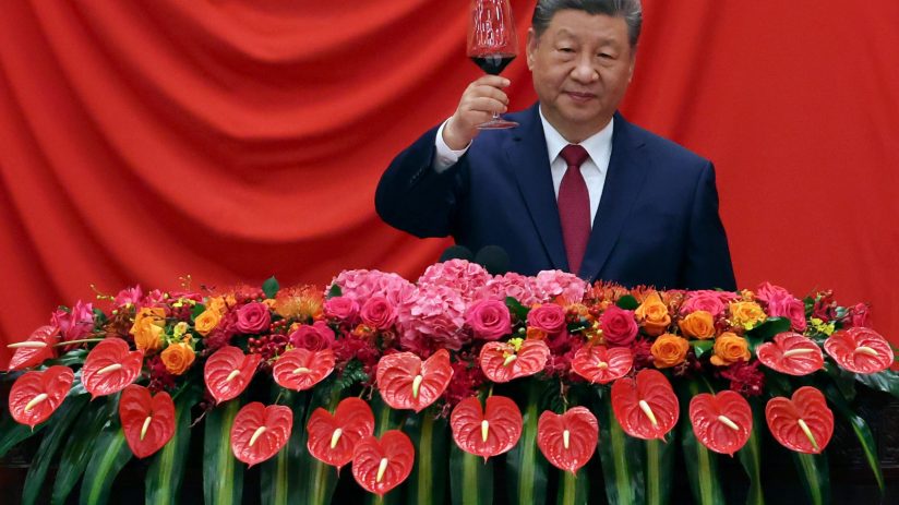 Xi Jinping in a dark suit and red tie, raising a glass of red wine, with a floral arrangement below him.