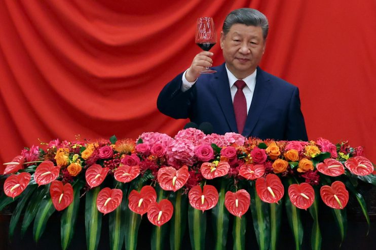 Xi Jinping in a dark suit and red tie, raising a glass of red wine, with a floral arrangement below him.