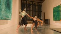 Two performers in an ornate room; one wears a horned mask, the other is in a crouch, holding hands.