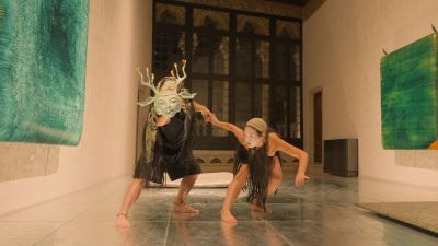 Two performers in an ornate room; one wears a horned mask, the other is in a crouch, holding hands.