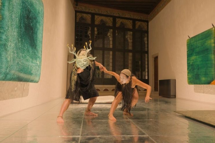 Two performers in an ornate room; one wears a horned mask, the other is in a crouch, holding hands.