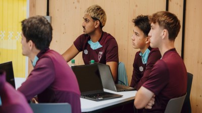 Young men in West Ham United shirts watching a presentation with laptops on a table in a modern room.