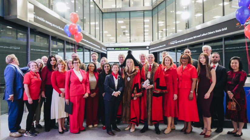 Group of people, some in red attire, with Lord Mayor at London Stock Exchange, FTSE 100 ticker visible.
