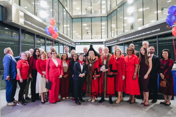 Group of people, some in red attire, with Lord Mayor at London Stock Exchange, FTSE 100 ticker visible.