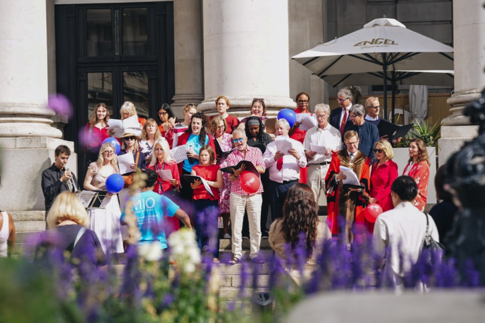 Mixed group of people singing outdoors on steps, some holding sheet music and balloons, with pillars and cafe umbrella in ...
