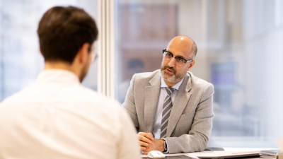 Businessman in glasses and suit jacket talks to another man across a desk in a modern office.