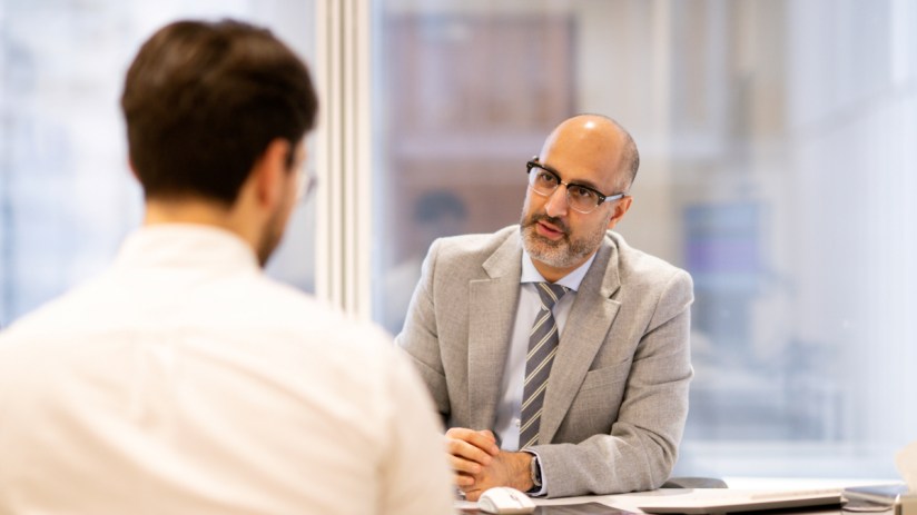 Businessman in glasses and suit jacket talks to another man across a desk in a modern office.
