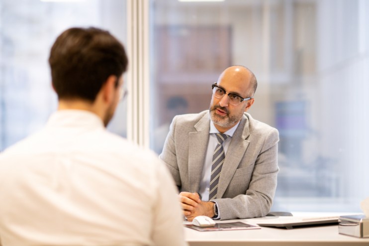 Businessman in glasses and suit jacket talks to another man across a desk in a modern office.