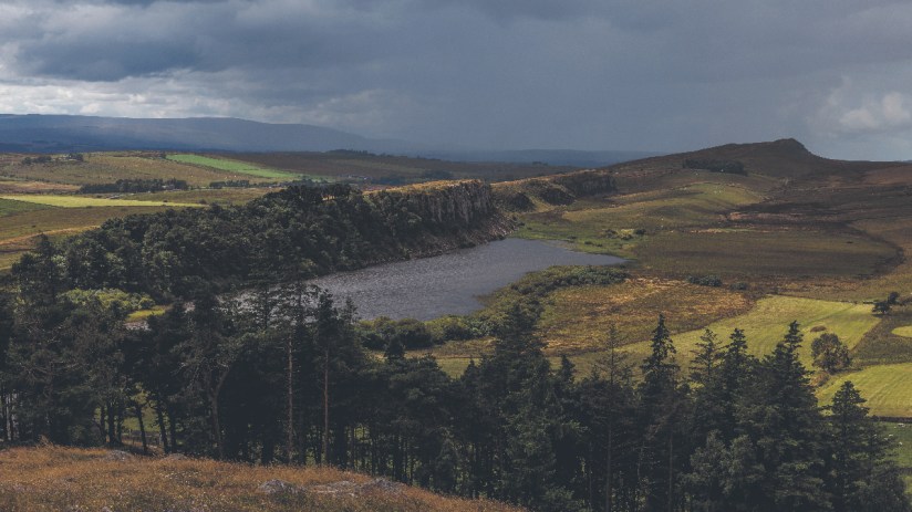 Dramatic Northumberland landscape with a dark lake, rugged cliffs, green fields, and a cloudy sky.