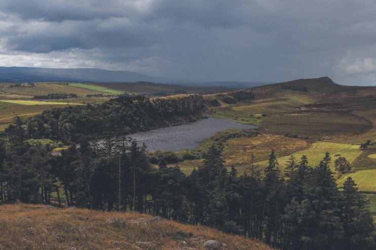 Dramatic Northumberland landscape with a dark lake, rugged cliffs, green fields, and a cloudy sky.
