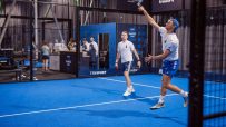Two men playing padel on a blue court, one serving with a racket overhead, another ready to receive.
