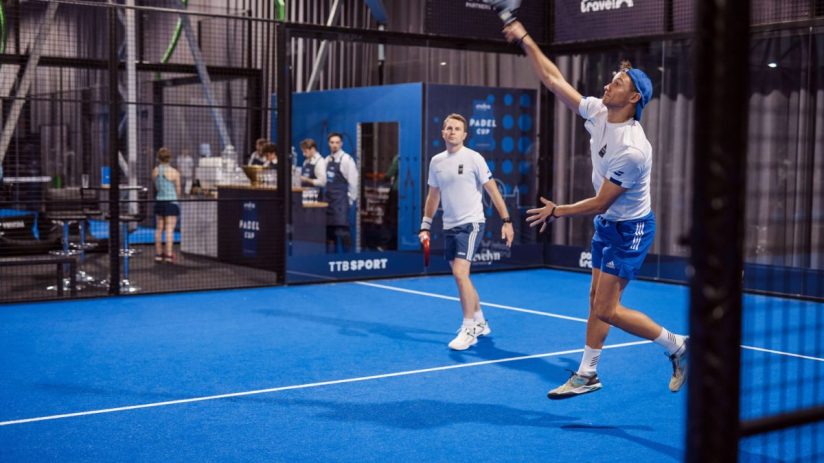 Two men playing padel on a blue court, one serving with a racket overhead, another ready to receive.