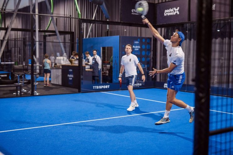 Two men playing padel on a blue court, one serving with a racket overhead, another ready to receive.