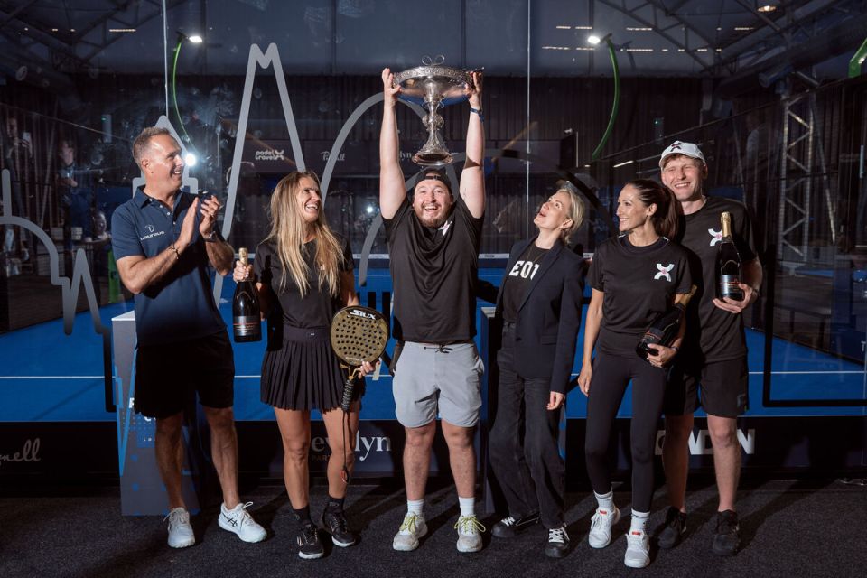 Six people celebrating on a padel court, man in center holding trophy, others with champagne bottles.