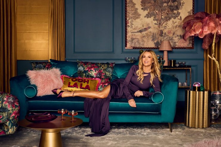 Amanda Holden reclining on a teal velvet sofa in a luxurious, richly decorated living room