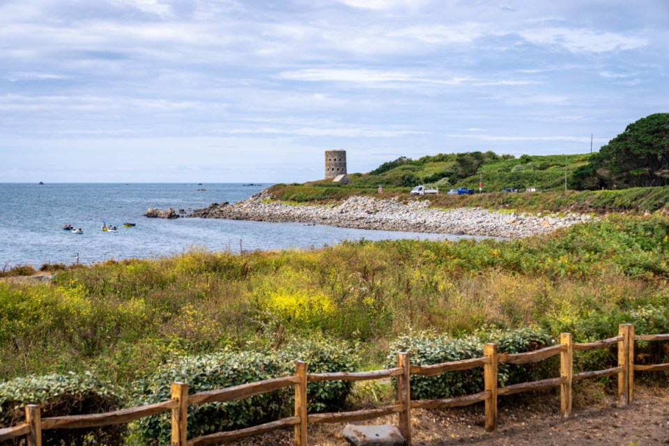 Guernsey coastline view with a historic Martello tower overlooking a rocky bay and distant kayaks, lush coastal vegetation...
