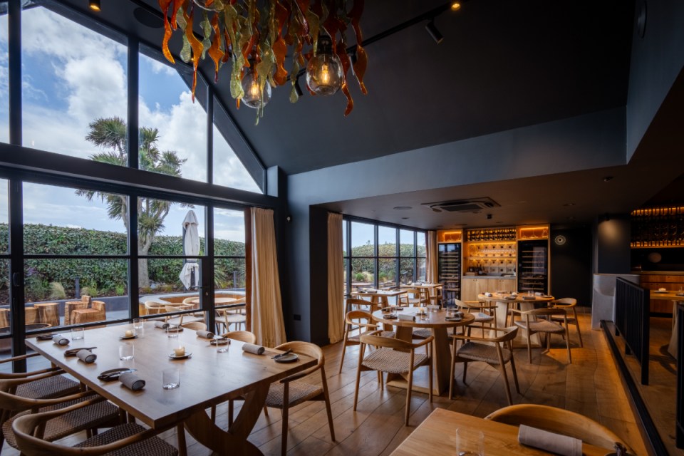 Modern restaurant interior with floor-to-ceiling windows, wooden tables, woven chairs, and an ornate chandelier.