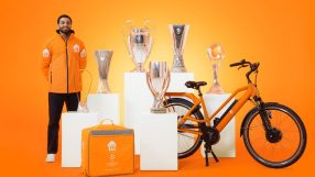Delivery driver in orange jacket with UEFA Champions League trophies, delivery bag, and orange bicycle.