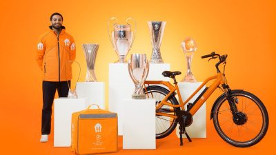 Delivery driver in orange jacket with UEFA Champions League trophies, delivery bag, and orange bicycle.