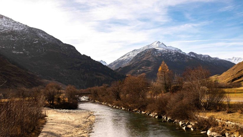 Andermatt Alps landscape in autumn with a river, snow-capped mountains, and golden-leafed trees under a blue sky.
