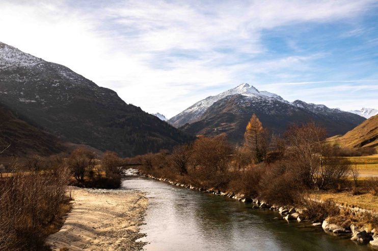 Andermatt Alps landscape in autumn with a river, snow-capped mountains, and golden-leafed trees under a blue sky.
