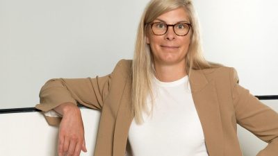 Angela Tangas, blonde woman in glasses and a tan blazer, leans casually on a white surface.