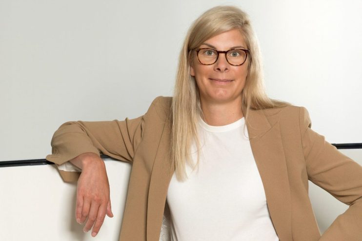 Angela Tangas, blonde woman in glasses and a tan blazer, leans casually on a white surface.