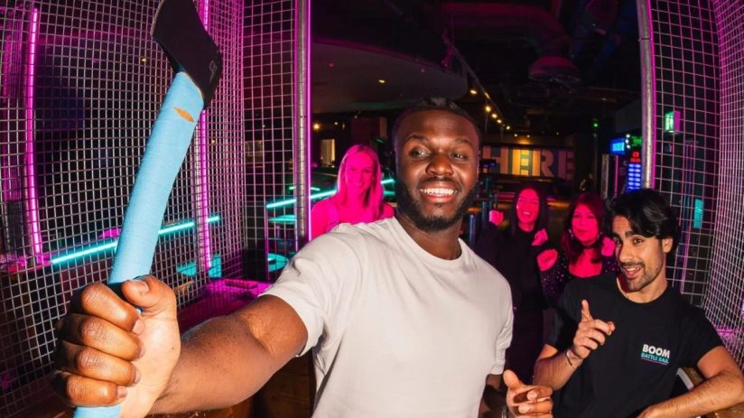 Smiling man holding a blue-handled axe in an axe-throwing lane with friends and neon lights