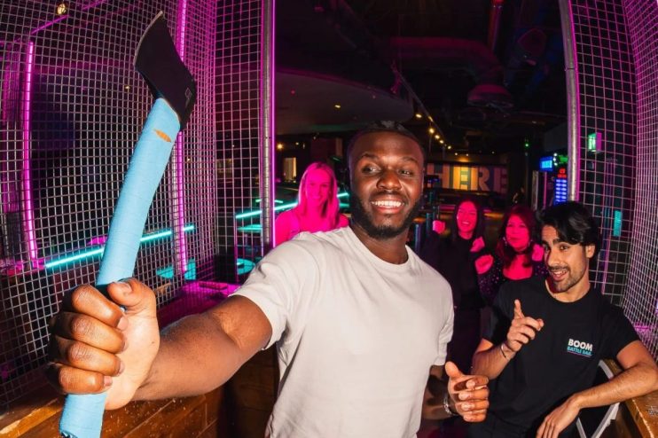 Smiling man holding a blue-handled axe in an axe-throwing lane with friends and neon lights