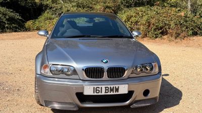 Front view of a silver BMW CSL parked on a dirt road with trees in the background.
