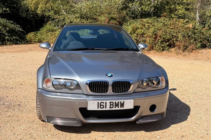 Front view of a silver BMW CSL parked on a dirt road with trees in the background.