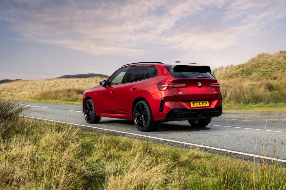 Red BMW X3 SUV on a winding road with grassy hills and a cloudy sky.