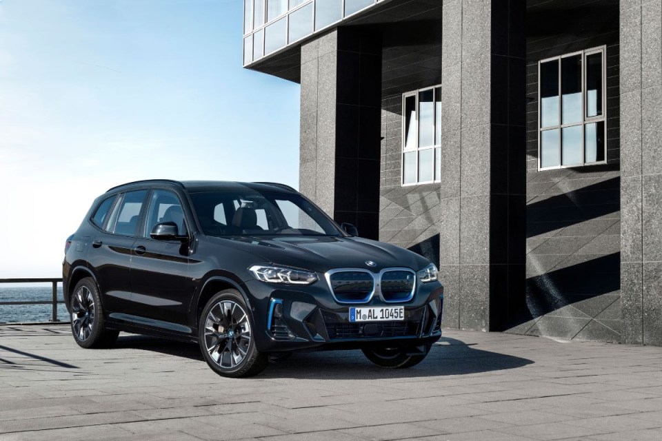 Black BMW iX3 electric SUV parked on a paved surface next to a modern building, with water in the background.