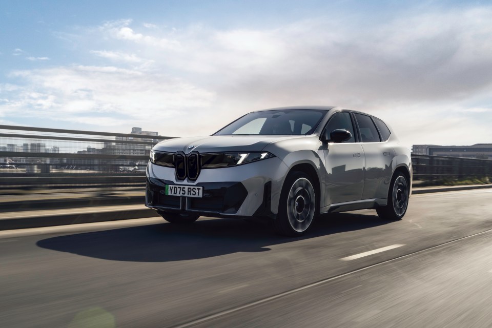 Silver BMW iX3 electric SUV driving on a highway with blurred background infrastructure and sky.
