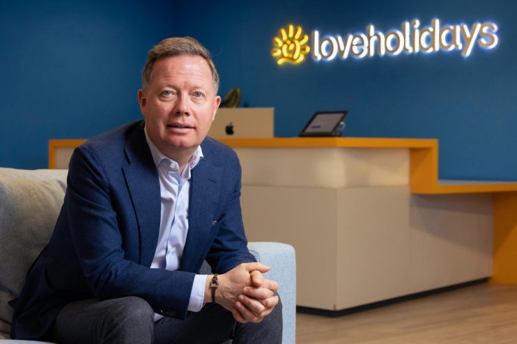 Man in suit seated in office with Loveholidays logo, representing business, travel, and tourism.